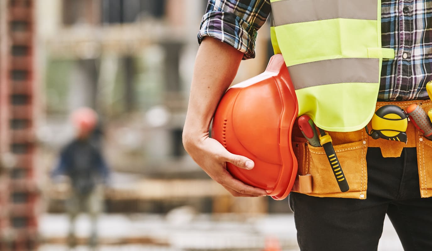 An image of a construction worker holding a helmet while also wearing a construction site vest and toolbelt.